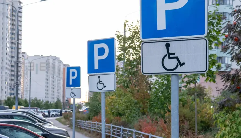 Znaki informują, komu przysługuje karta parkingowa. Na parkingu stoją samochody, a w tle widać budynki mieszkalne.