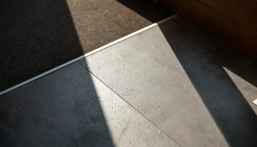 Un tappeto scuro incontra un moderno pavimento flottante grigio, illuminato da un raggio di sole diagonale.