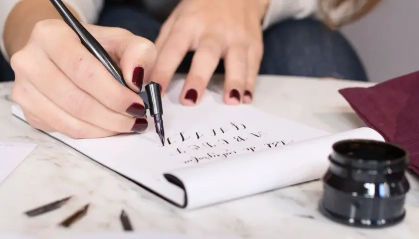 Manos practicando lettering con pluma y tinta, creando letras cursivas en un cuaderno.