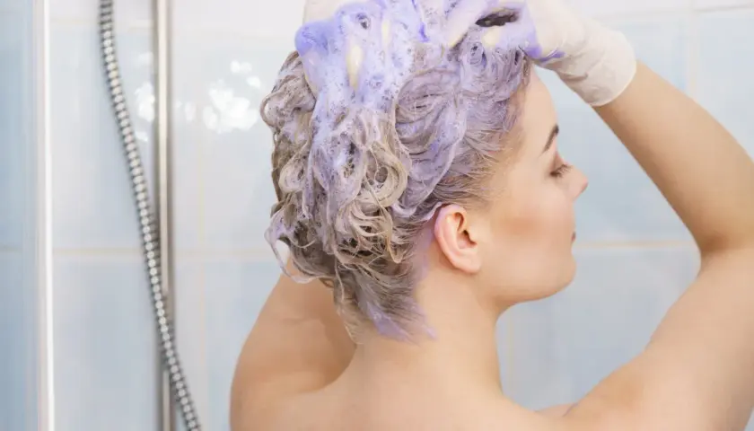 Kobieta w rękawiczkach nakłada fioletowy szampon na blond włosy pod prysznicem.