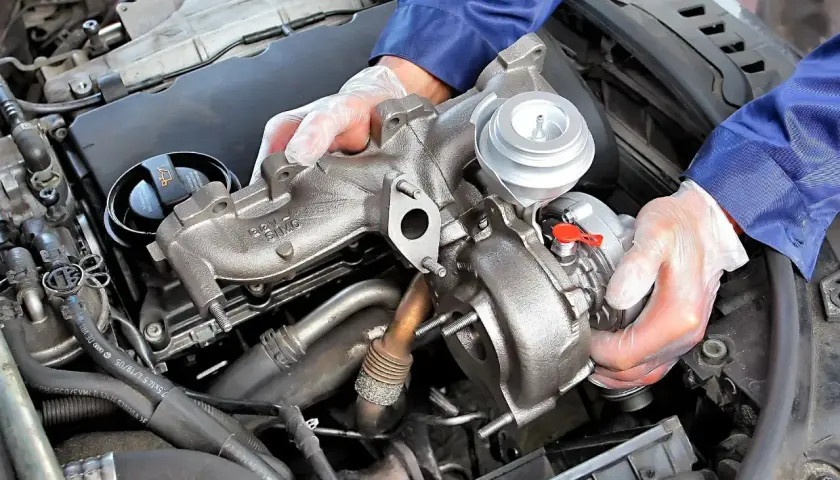 Mechanik w rękawiczkach trzyma turbosprężarkę. Uszkodzony czujnik doładowania turbiny objawy mogą być różne, ale często wiążą się z utratą mocy.