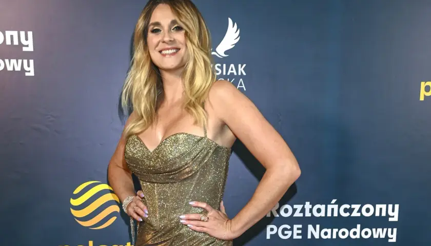 Kobieta w złotej sukience z cekinami, uśmiechnięta, na tle granatowej ścianki z logo Polsat.
