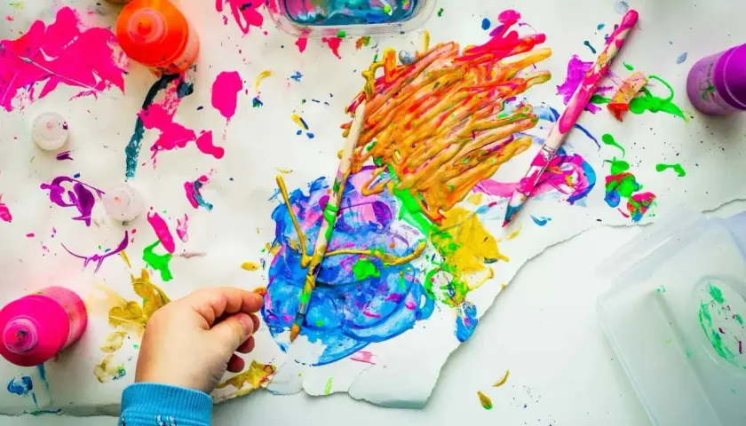 Kinderhand malt mit bunten Farben auf Papier, umgeben von Farbtöpfen und Pinseln.