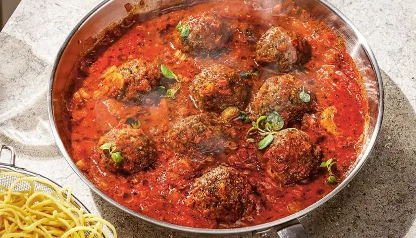 Frikadeller i tomatsås som puttrar i en panna, med en skål spaghetti bredvid.