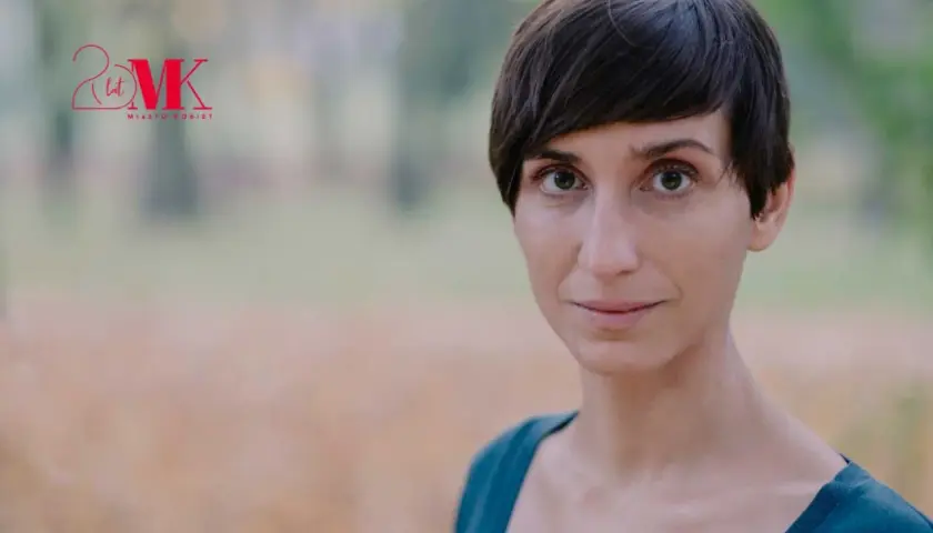 Kobieta po przejściach, z krótkimi włosami i przenikliwym spojrzeniem, uśmiecha się lekko. W tle logo "20 lat Miasto Kobiet".