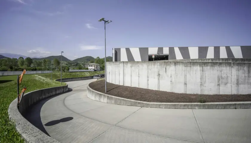 Ampia rampa in cemento fai da te, curva dolcemente verso un edificio moderno con facciata a strisce bianche e grigie, circondata da verde e colline.