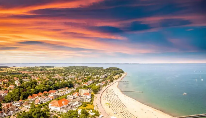 Ein perfekter tagesausflug ostsee: Sonnenuntergang über der Ostsee, belebter Strand mit Liegen und ein Küstenort mit roten Dächern.