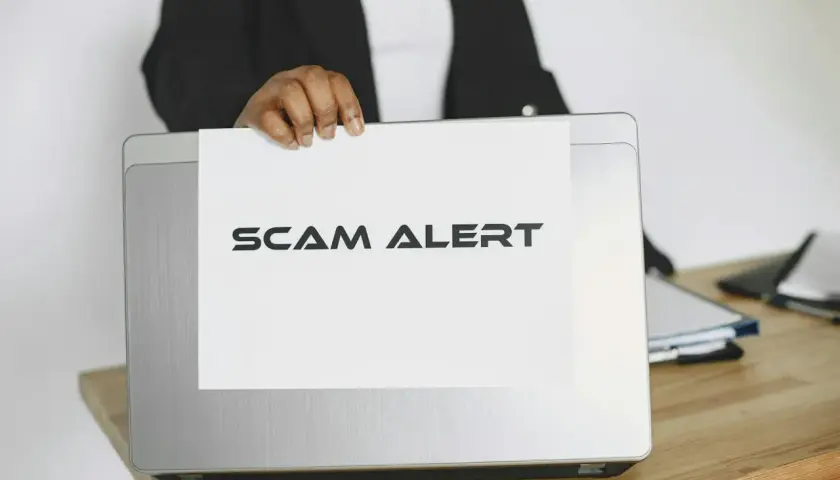 Ostrzeżenie przed oszustwami na Vinted. Ktoś trzyma kartkę z napisem "SCAM ALERT" nad laptopem.