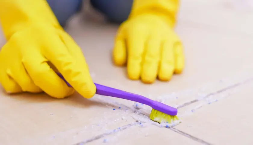 Hände in gelben Handschuhen reinigen mit einer lila Zahnbürste Fliesen und Fugen.