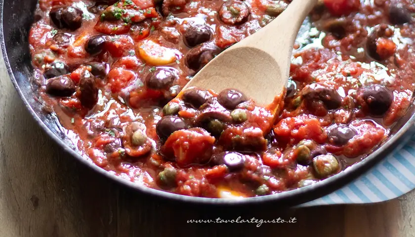 Un cucchiaio di legno mescola un sugo ricco e saporito, pieno di pomodori, olive nere e capperi. È la base perfetta per una pasta alla puttanesca.