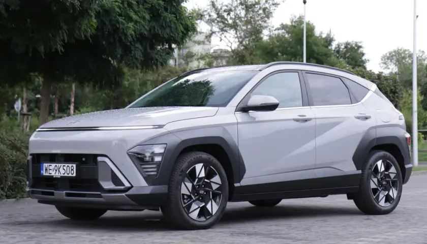 Nowy Hyundai Kona Hybrid w kolorze jasnoszarym, zaparkowany na brukowanym placu, otoczony zielenią.