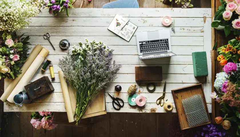 Kwiaty, laptop i akcesoria florystyczne, jak nożyczki, wstążki i sznurek, tworzą inspirujący warsztat. Widać też materiały do pakowania, przypominające **faszynę**.