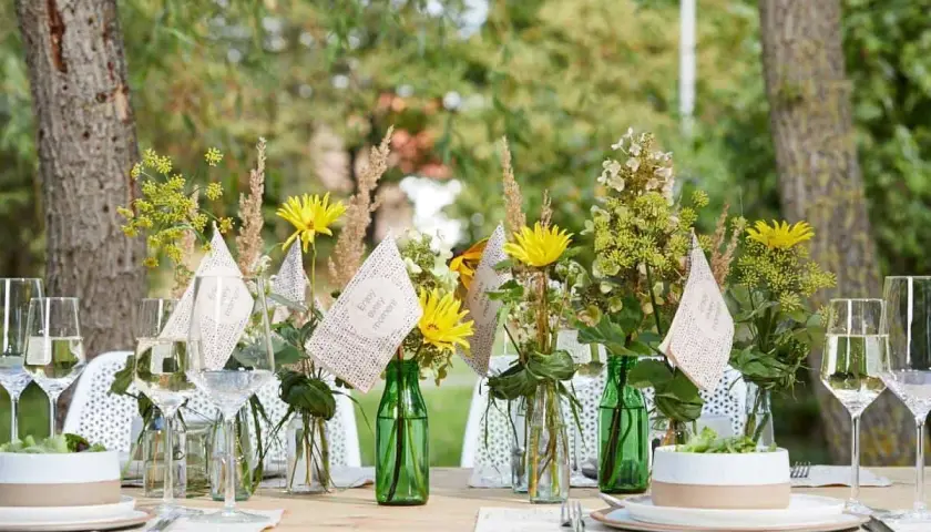 Einfache Tischdeko Sommer: Gelbe Blumen in grünen Flaschen, Weingläser und kleine Fähnchen mit der Aufschrift "Enjoy every moment" auf einem Holztisch im Freien.