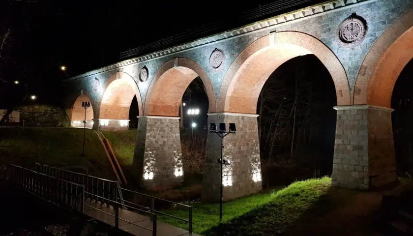 Nocna iluminacja kamiennego wiaduktu z ceglanymi łukami. Widać fragment mostku i drzewa. To piękna bytów trasa.