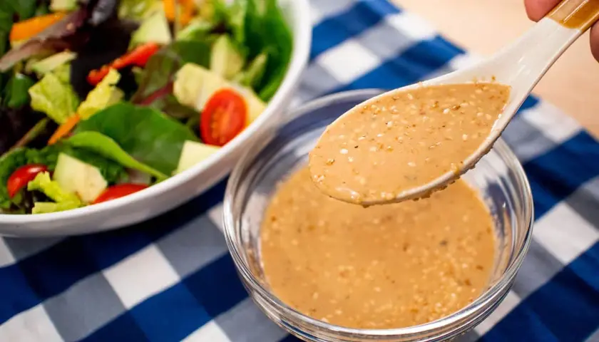 A spoon lifts creamy roasted sesame dressing from a bowl, ready to dress a fresh salad with avocado and tomatoes.