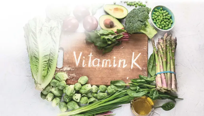 Grünes Gemüse wie Spinat, Brokkoli und Rosenkohl, das auf einem Holzbrett mit der Aufschrift "Vitamin K" arrangiert ist, zeigt, was Vitamin K ist.
