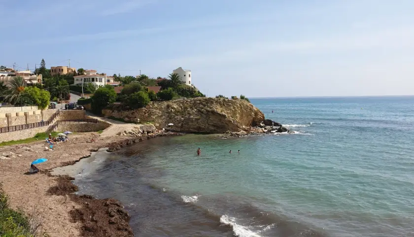 La cala Coveta, con su playa de arena y rocas, invita a disfrutar del mar Mediterráneo. Casas blancas coronan la colina.