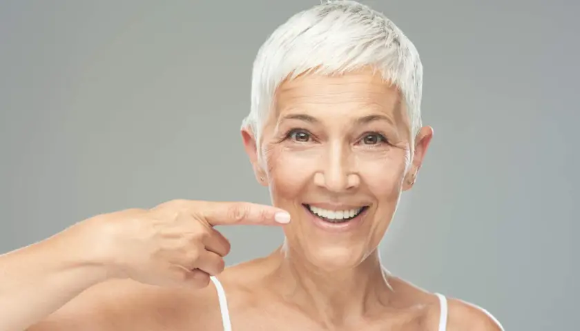 Donna sorridente indica il suo sorriso, un esempio di come rinforzare i denti a cinquant'anni con un sorriso smagliante.
