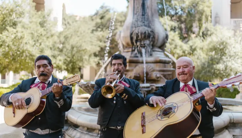 Trzech muzyków mariachi gra na gitarach i trąbce przy fontannie.