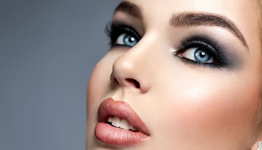 Niebieskie oczy z mocnym makijażem typu smokey eyes, podkreślone długimi rzęsami i ciemnymi cieniami. Usta w odcieniu nude.