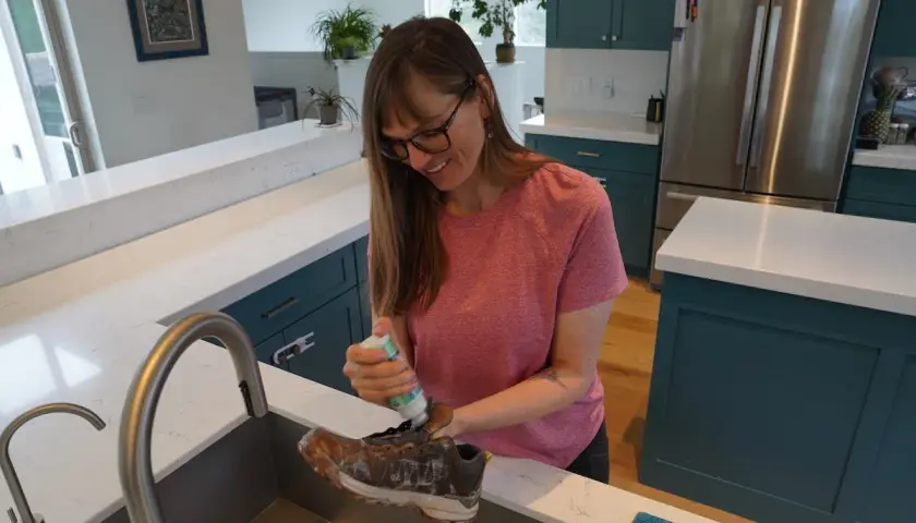 A woman cleans a dirty boot in a kitchen sink, preparing it for the washer.