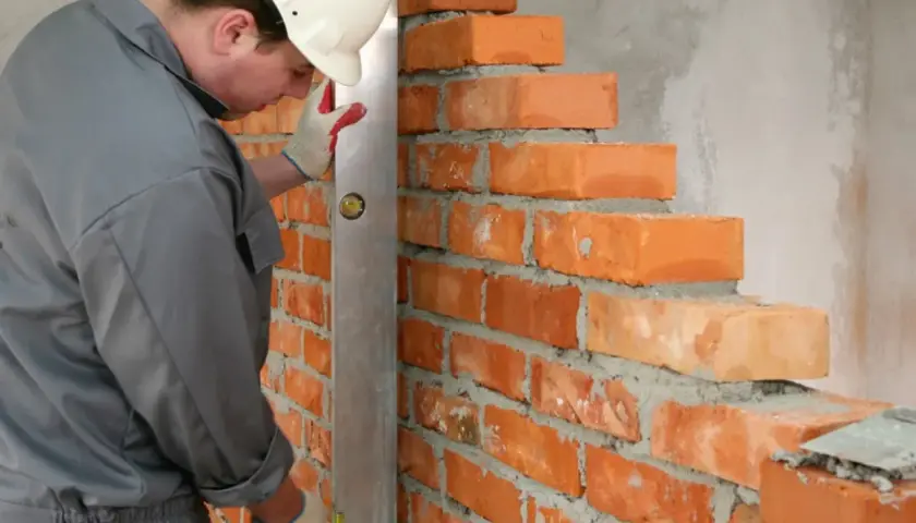 Murarz w kasku sprawdza pion muru, układając cegły. Dokładne wymiary cegły są kluczowe dla równego muru.