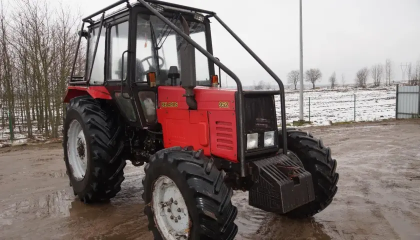 Czerwony ciągnik Belarus 952 z kabiną i osłoną, stojący na błotnistym podwórzu. W tle pole z resztkami śniegu.