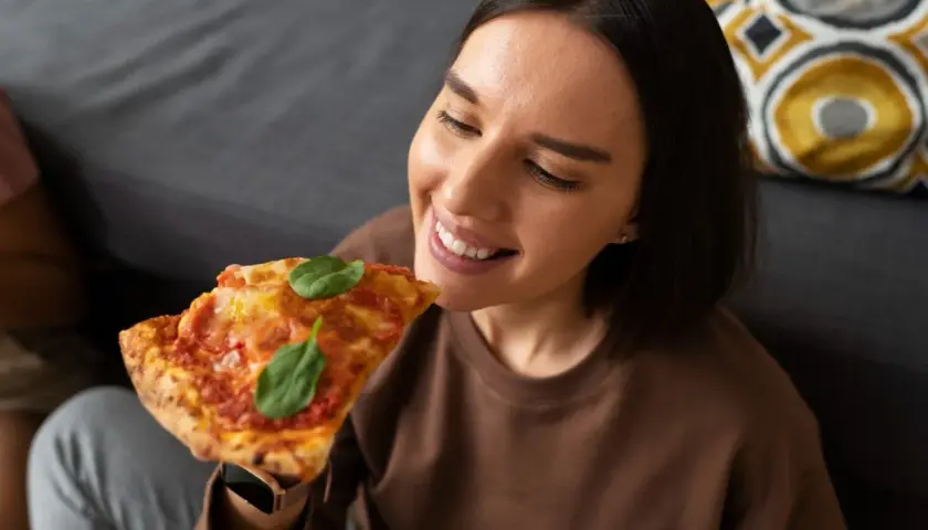 Uśmiechnięta kobieta delektuje się kawałkiem pizzy, zastanawiając się, jak podgrzewać pizzę, by była idealna.