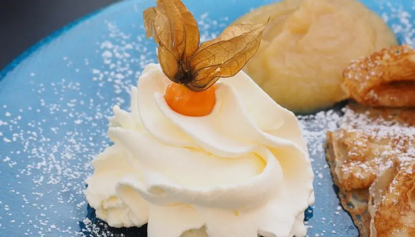Panna montata soffice con physalis, su un piatto blu cosparso di zucchero a velo, accanto a un dolce che ricorda il pan di spagna.