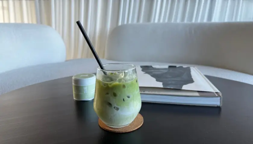 Un verre de matcha vert glacé, avec une paille noire, sur un sous-verre en liège. Une boîte de matcha est à gauche, un livre à droite.