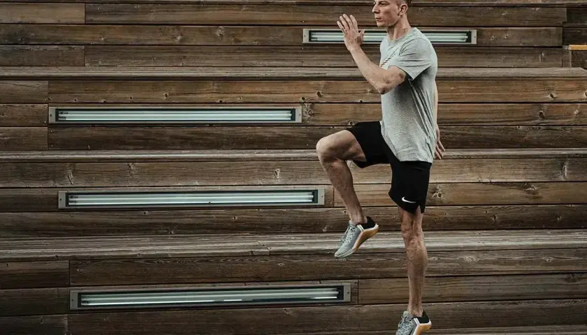 Mężczyzna w szarej koszulce i czarnych spodenkach wykonuje dynamiczne ćwiczenie, podnosząc kolano. Intensywny trening HIIT na drewnianych schodach.
