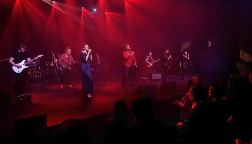 Zespół muzyczny na scenie, oświetlony czerwonymi światłami. Wokalistka w granatowej sukni, obok niej gitarzyści i wokalista w czerwonej koszuli. W tle widać perkusję i innych muzyków. Publiczność siedzi w ciemności. To jakby **sufler** podpowiadał im k...
