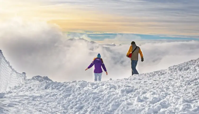 Dwie osoby wędrują po ośnieżonym szczycie. W oddali widać góry i chmury. Czyżby szukali odpowiedzi na pytanie: jakie raki w Tatry?