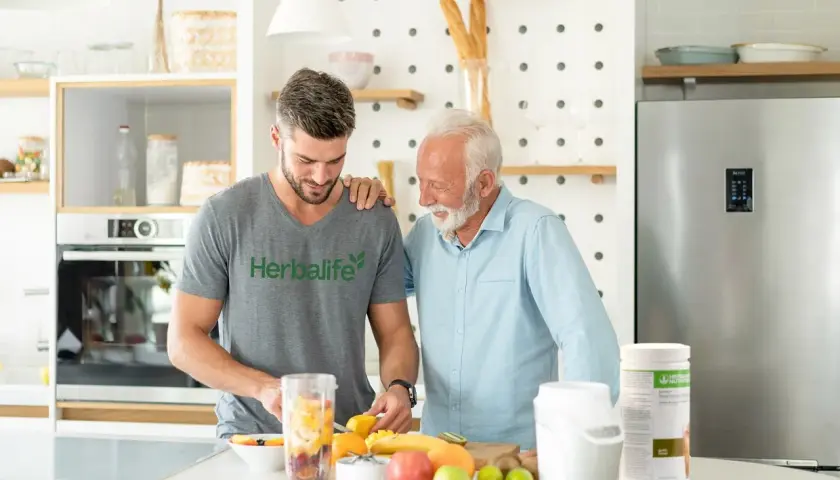 Dwóch mężczyzn przygotowuje smoothie z owocami. Młodszy w koszulce Herbalife, starszy w błękitnej koszuli. Dyskutują, ile odżywki białkowej dziennie dodać do koktajlu.