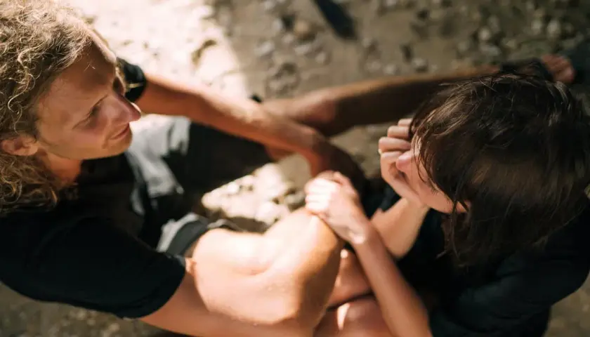 Un uomo con lunghi capelli biondi guarda una donna che gli tocca il braccio. Sono seduti sulla sabbia.