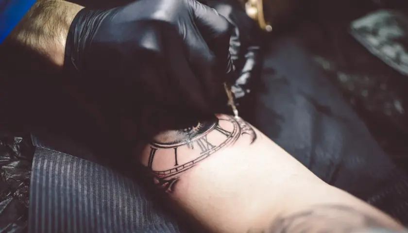 Artysta w czarnej rękawiczce tworzy tatuaż zegara, używając techniki whip shading do dodania głębi i cieni.