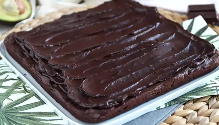 Puszysty brownie z awokado, polane błyszczącą polewą czekoladową. Obok połówka awokado i kawałek gorzkiej czekolady.