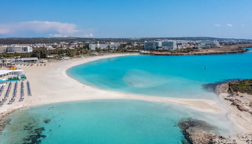 Piękna plaża Ayia Napa z turkusową wodą i białym piaskiem, otoczona hotelami i zielenią.