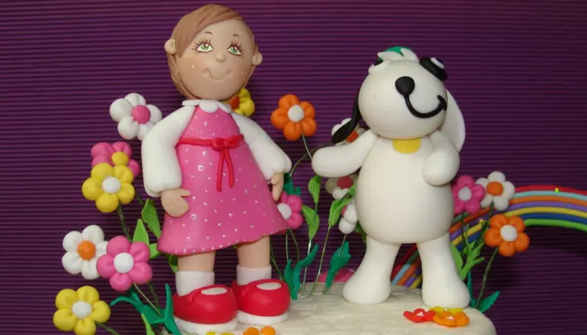 Figuras de porcelana fría: niña con vestido rosa y perro blanco rodeados de flores y un arcoíris.