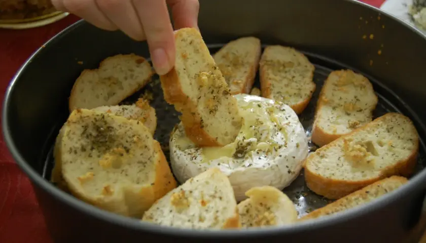 Ciepły serek camembert w okrągłej formie, otoczony kromkami pieczywa z ziołami. Ktoś zanurza kawałek chleba w rozpływającym się serze.