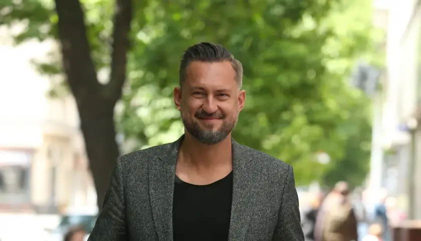 Marcin Prokop w szarej marynarce i czarnej koszulce, uśmiecha się na tle zieleni.