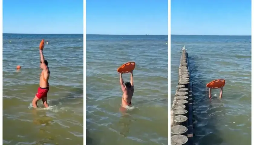 Ratownik z kołem ratunkowym w wodzie, obok drewnianego falochronu.