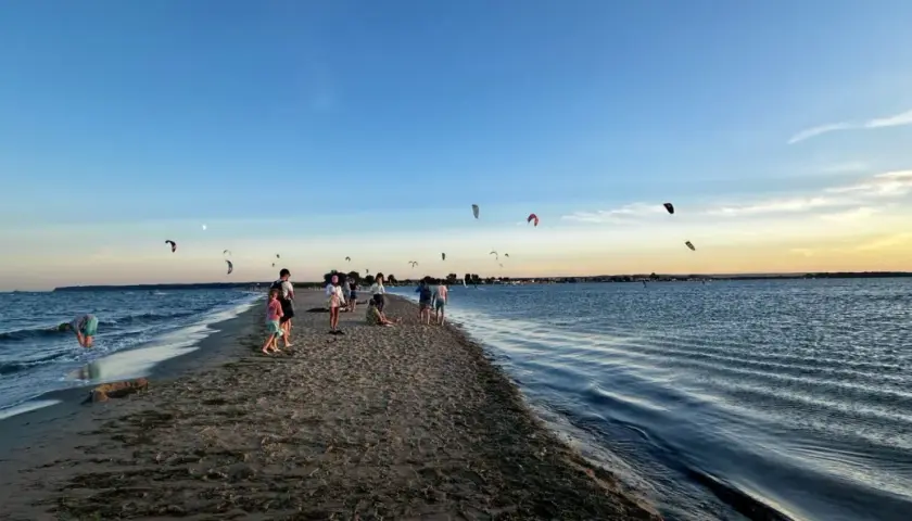 Ludzie spacerują po piaszczystej mierzei, podczas gdy na niebie widać liczne latawce do kitesurfingu.