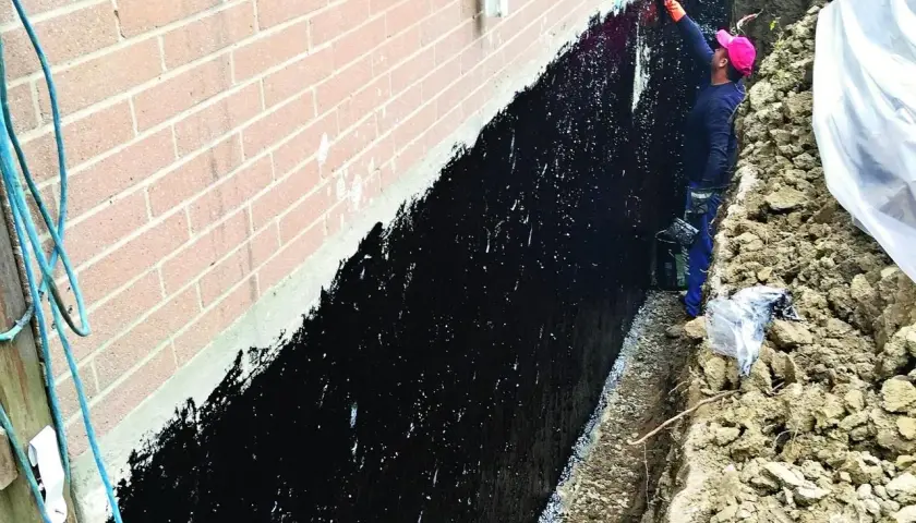 Pracownik zabezpiecza ławy fundamentowe czarną masą izolacyjną.
