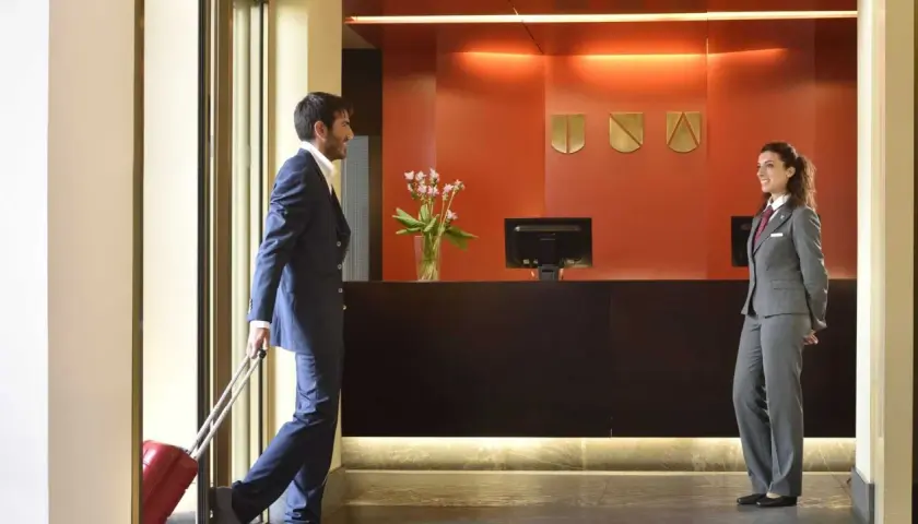 Uomo con valigia rossa alla reception, pronto per l'early check-in. La receptionist sorride accogliendolo.