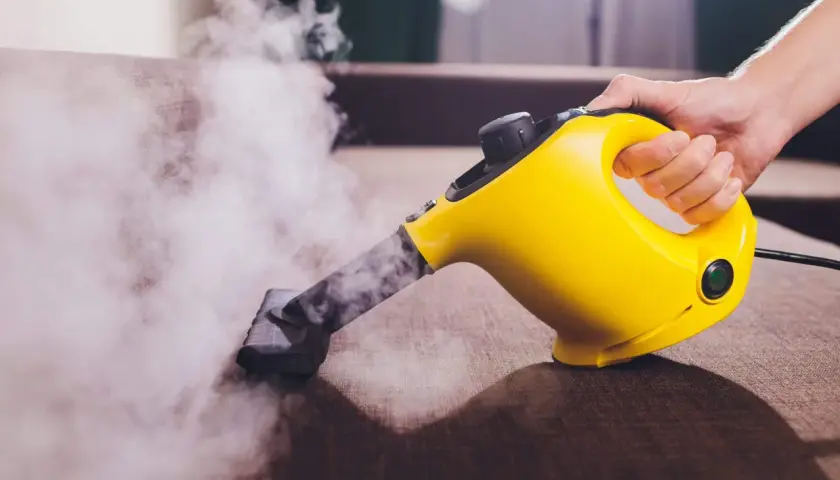 Mano sujetando una limpiadora a vapor amarilla, desprendiendo vapor sobre un sofá marrón.