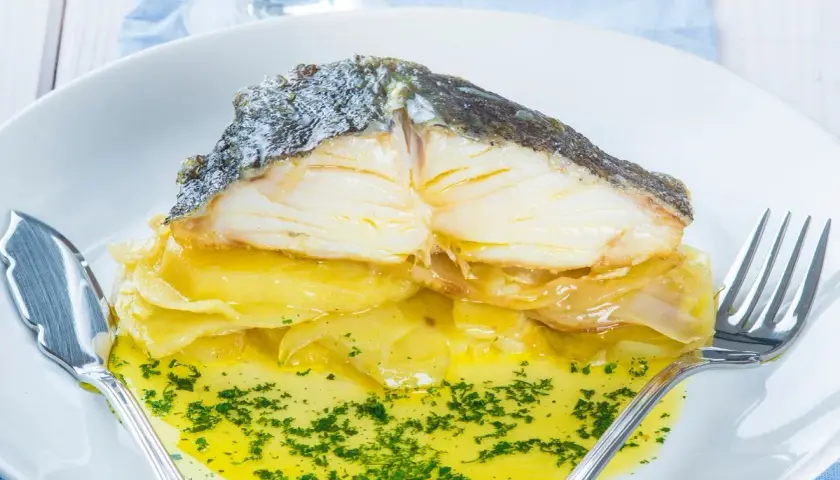 Merluza al horno con patatas, jugosa y dorada, servida sobre una cama de patatas y cebolla caramelizada, bañada en aceite de oliva y perejil picado.