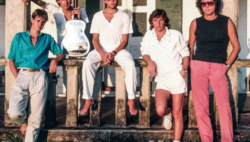 Członkowie Dire Straits pozują na tle budynku. Jeden z nich trzyma gitarę.