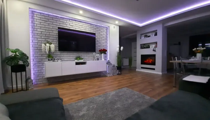 Nowoczesny salon z ceglaną ścianą i telewizorem, pięknie podświetloną fioletowym światłem. Świetny pomysł na podświetlenie ściany!