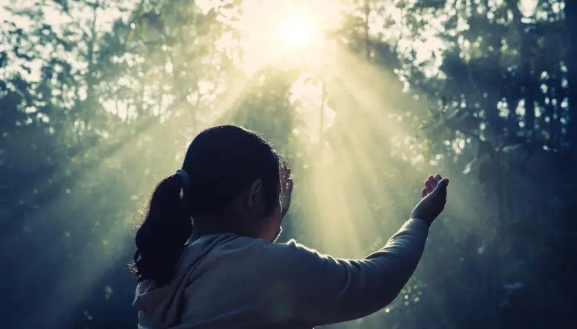 Una niña con coleta, con las manos alzadas, parece soñar con espíritus mientras la luz del sol atraviesa los árboles.